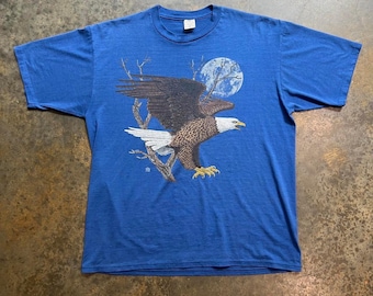 May include: A blue t-shirt featuring a graphic of a bald eagle flying in front of a full moon.