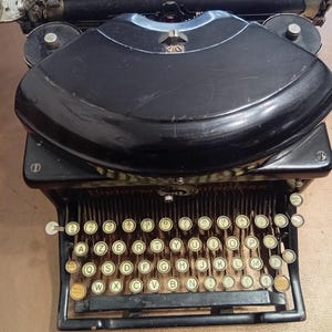 Puede incluir: Una máquina de escribir vintage Underwood Noiseless negra con un teclado QWERTY estándar y teclas amarillas. La máquina de escribir tiene un cuerpo de metal negro y una parte superior curva.