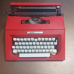 Peut inclure: Une machine à écrire Olivetti COLLEGE rouge avec un clavier blanc et des touches noires. La machine à écrire a un levier de retour chariot noir et une bobine de ruban noire.
