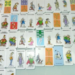 Peut inclure: Un ensemble de dominos blancs ornés d'illustrations de personnages de dessins animés colorés. Chaque domino présente un design unique, certains avec des points et d'autres avec des images complètes de personnages. Les dominos sont disposés de près.