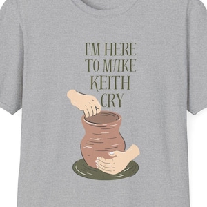 Puede incluir: Camiseta gris jaspeado con el texto "I'M HERE TO MAKE KEITH CRY" sobre una ilustración de manos modelando un jarrón de cerámica marrón. El diseño es minimalista, centrándose en el texto y la cerámica.