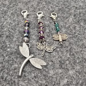 May include: Three dragonfly-themed charms with silver-tone clasps. One charm features a large, textured dragonfly pendant with a curved body and outstretched wings. The other two charms have beaded accents in purple, green, and clear colors.