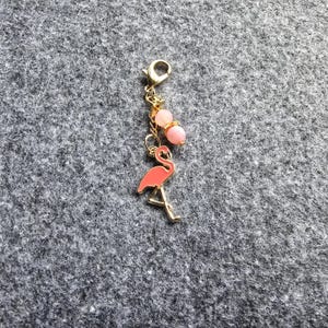 May include: A gold-toned metal charm featuring a pink flamingo pendant and two small, round, light pink beads. The charm is attached to a gold-toned lobster clasp by a short metal chain, creating a decorative accessory for bags or jewelry.