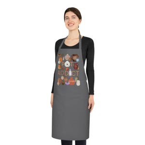 Może przedstawiać: Szary fartuch z napisem "POT ADDICT" i ilustracjami ceramiki. Fartuch jest noszony przez osobę w czarnej bluzce z długim rękawem.