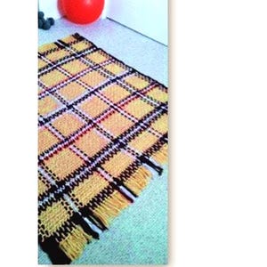 May include: A yellow and black plaid woven rug with a fringed edge. The rug has a repeating pattern of squares with red and white accents. A red ball and some blue berries are in the background.