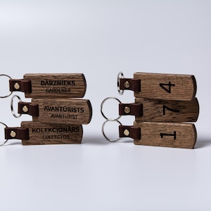 May include: Wooden keychains with metal rings and leather straps. Some keychains have the words "DĀRZNIEKS GARDENER", "AVANTŪRISTS AVANTURIST", and "KOLEKCIONĀRS COLLECTOR" printed on them. Other keychains have the numbers "1", "7", and "4" printed on them.