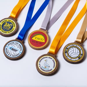 Op de afbeelding: Een verzameling houten medailles met kleurrijke linten. De medailles hebben verschillende ontwerpen, waaronder een taco, een volleybal en tekst zoals "1st Annual" en "Champions". De linten zijn in tinten geel, blauw en grijs.