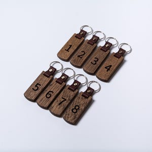 May include: Eight rectangular wooden keychains arranged in two rows. Each keychain features a dark brown leather strap attached with a brass rivet to a textured wood piece engraved with a black number from one to eight and a silver metal ring.