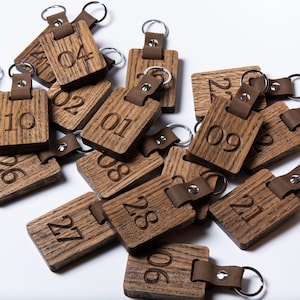 May include: A pile of wooden keychains with leather straps and metal rings. Each keychain has a number engraved on it, ranging from 01 to 28.