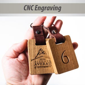 May include: Two wooden keychains with leather straps. One keychain is engraved with the text "2413m Rifugio Averau Dolomiti" and the other keychain is engraved with the number "6".