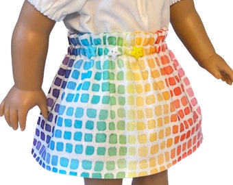 May include: A doll wearing a white short-sleeved top and a rainbow-colored skirt. The skirt features a grid pattern of squares in various colors, including purple, blue, green, yellow, and red. The skirt has a yellow star-shaped button and a white star-shaped button.