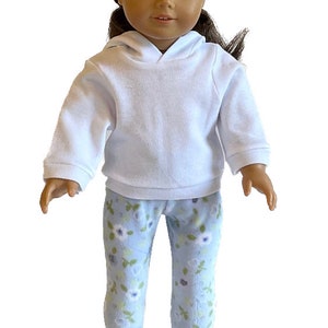 May include: A doll wearing a white hooded sweatshirt and light blue floral print pajama pants. The doll has brown hair and brown eyes. The doll is standing and facing forward.