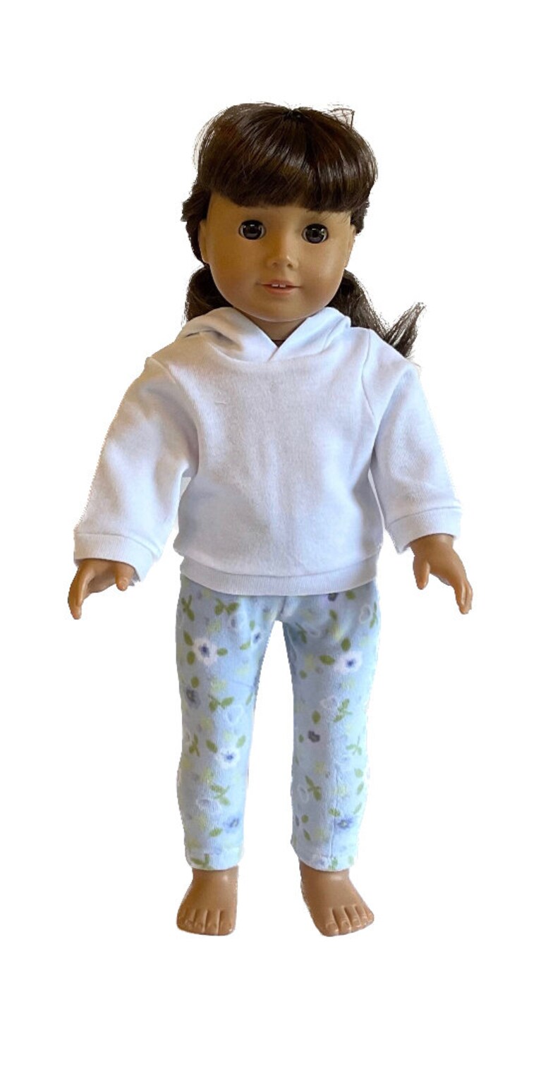 May include: A doll wearing a white hooded sweatshirt and light blue floral print pajama pants. The doll has brown hair and brown eyes. The doll is standing and facing forward.
