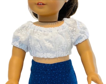 May include: A white, short-sleeved crop top with floral embroidery and a blue skirt with a polka dot pattern. The doll clothing set is designed for a doll.