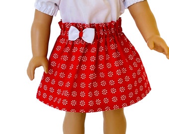 May include: A doll wearing a red skirt with a white daisy pattern and a white bow. The skirt has an elastic waistband. The doll is also wearing a white top with short sleeves.