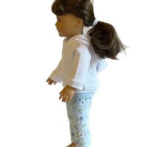 May include: A doll wearing a white hooded sweatshirt and light blue floral print pants. The doll has brown hair pulled back in a ponytail. The doll is barefoot.