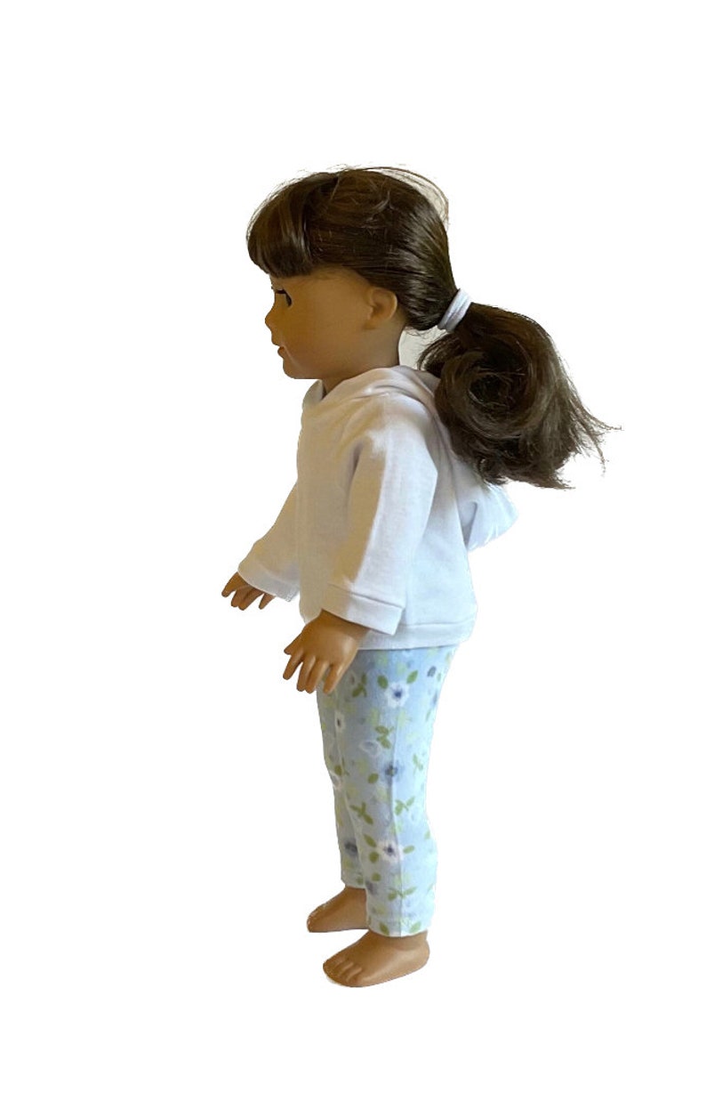 May include: A doll wearing a white hooded sweatshirt and light blue floral print pants. The doll has brown hair pulled back in a ponytail. The doll is barefoot.