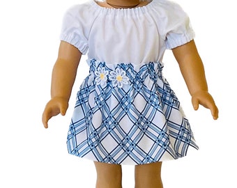 May include: A doll wearing a white short-sleeved top and a blue and white plaid skirt. The skirt has a gathered waistline and is decorated with two white daisy appliqués with yellow centers. The outfit is designed for a doll.