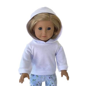 May include: A doll wearing a white hooded sweatshirt and blue floral print pants. The doll has blonde hair and blue eyes. The sweatshirt has long sleeves and a ribbed hem. The pants have a light blue background with a floral pattern.