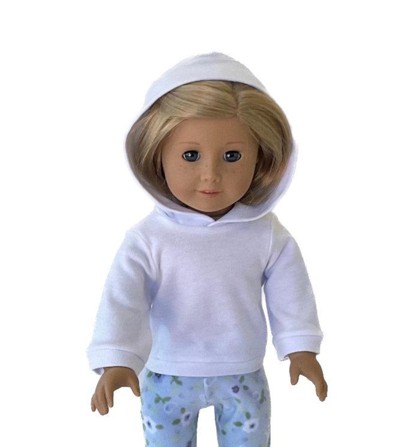 May include: A doll wearing a white hooded sweatshirt and blue floral print pants. The doll has blonde hair and blue eyes. The sweatshirt has long sleeves and a ribbed hem. The pants have a light blue background with a floral pattern.
