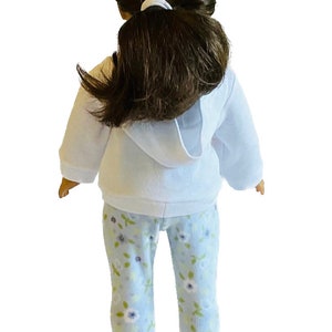 May include: A doll wearing a white hooded sweatshirt and light blue floral print pajama pants. The doll has dark brown hair pulled back in a ponytail. The pajamas have a pattern of white and purple flowers.