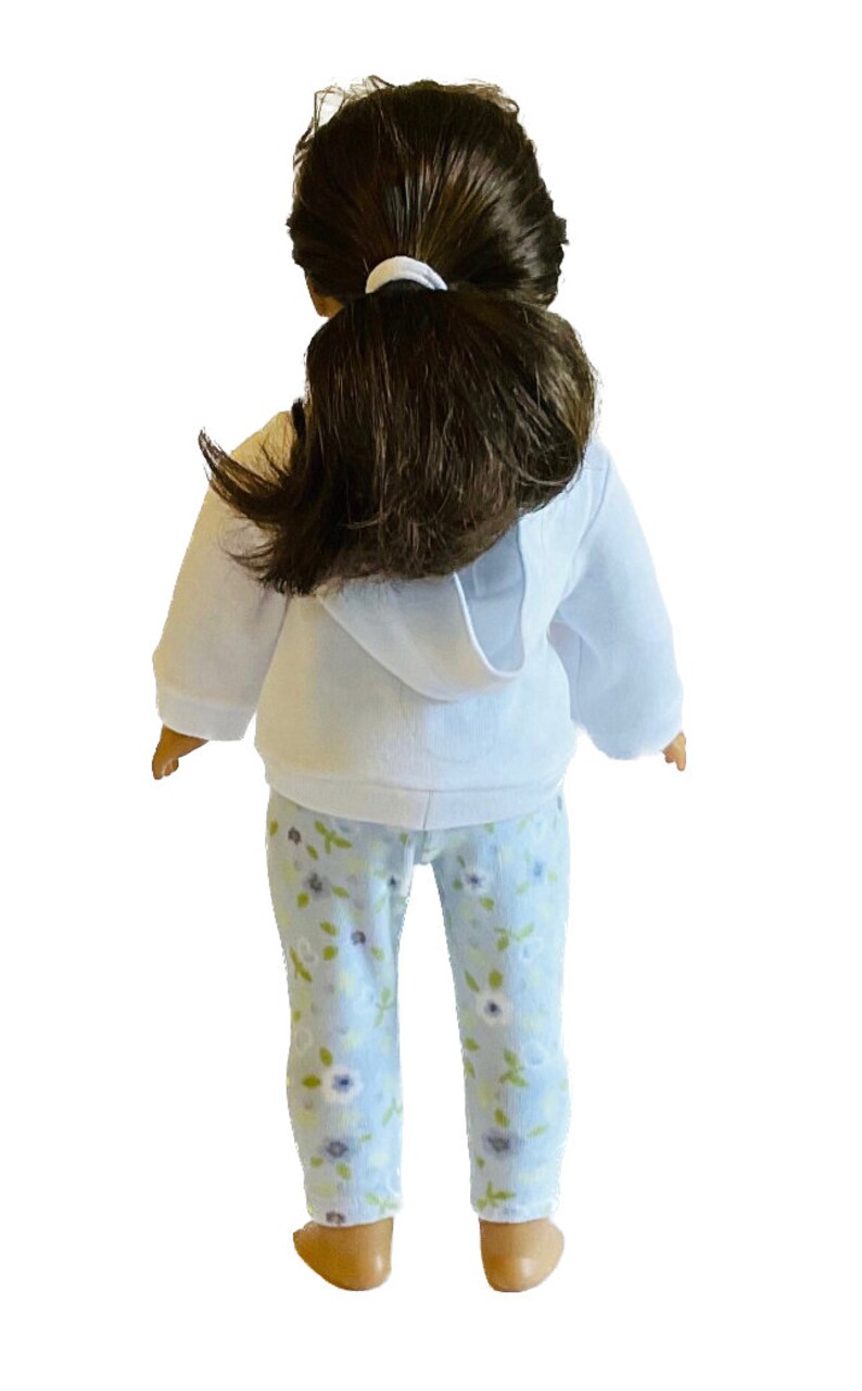 May include: A doll wearing a white hooded sweatshirt and light blue floral print pajama pants. The doll has dark brown hair pulled back in a ponytail. The pajamas have a pattern of white and purple flowers.
