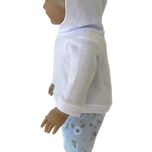 May include: A doll wearing a white hooded sweatshirt and light blue floral print pajama pants. The doll's outfit is designed for comfort and play, perfect for imaginative adventures. The doll is standing.