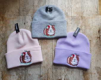 May include: Three knit beanies with different colored embroidery of a guinea pig wearing a flower crown. The beanies are gray, pink, and purple.