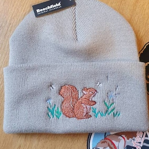 May include: Grey knitted beanie with an embroidered squirrel design. The beanie has a fold-over cuff and a black tag that reads "Beechfield Original Headwear".