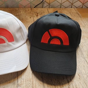 Könnte beinhalten: Zwei Baseballkappen, eine weiße und eine schwarze, mit einem roten Logo auf der Vorderseite. Das Logo ist eine stilisierte geometrische Form.