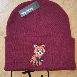 Può includere: Un berretto bordeaux con risvolto e un panda rosso ricamato sul davanti. Il panda rosso ha segni arancioni e neri. Un'etichetta nera con la scritta "Beechfield ORIGINAL HEADWEAR" è attaccata.