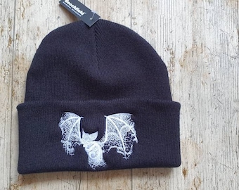 Pode incluir: Um gorro de malha preto com um morcego branco bordado apresentando padrões de asas intrincados semelhantes a renda. O gorro inclui uma dobra na barra e uma pequena etiqueta preta no topo com o texto branco Beechfield Original Headwear.