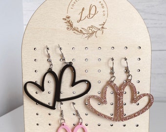May include: A wooden earring display stand with three pairs of heart-shaped earrings. The earrings are black, pink, and sparkly gold.