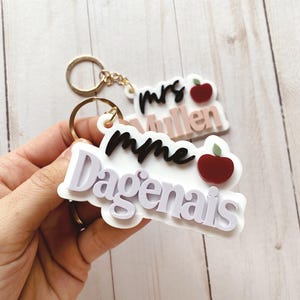Puede incluir: Dos llaveros con temática de profesor. Un llavero dice "mrs Mullen" en negro y rosa con una manzana roja. El otro llavero dice "mme Dagenais" en blanco y negro con una manzana roja. Ambos llaveros tienen llaveros dorados.
