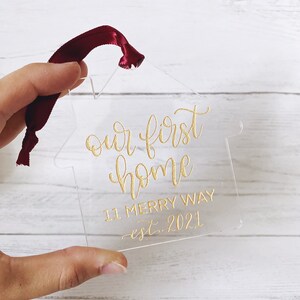 May include: Clear acrylic house-shaped ornament with gold script text reading "our first home", "11 Merry Way", and "est. 2021". A burgundy ribbon is attached for hanging. A housewarming gift.