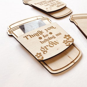 Puede incluir: Etiqueta de regalo de madera en forma de taza de caf&eacute; con el texto "Gracias por ayudarme a crecer" y un peque&ntilde;o dise&ntilde;o floral. La etiqueta est&aacute; dise&ntilde;ada para contener una tarjeta de regalo o una peque&ntilde;a nota.