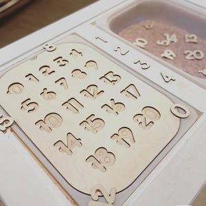May include: A light-colored wooden board featuring laser-cut numbers from 0 to 20, with matching wooden number pieces placed on top and inside a white tray. A small white label in the corner displays the text CHOKING HAZARD 3+.