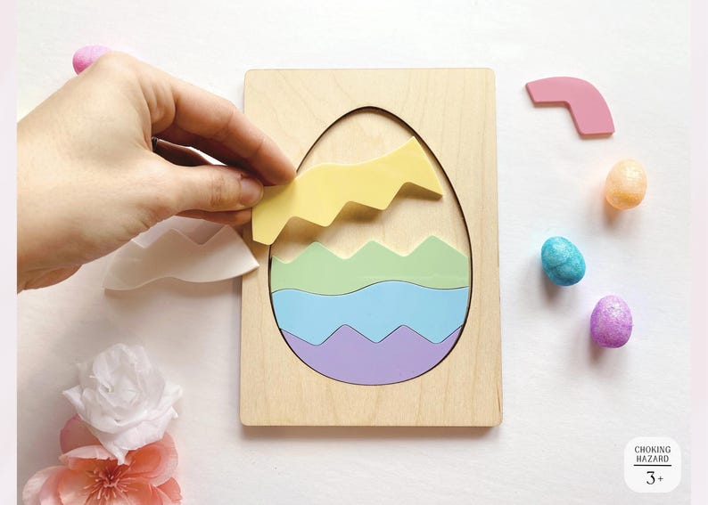 May include: A wooden egg-shaped puzzle board with interlocking pieces in pastel yellow, green, blue, and purple. The puzzle features a cutout egg shape, with additional loose pieces in white and pink, alongside small decorative eggs and a flower.