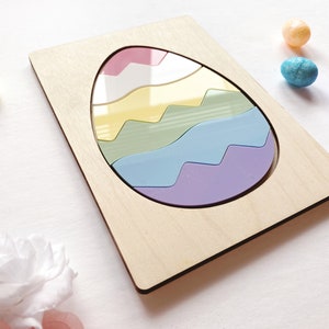 May include: A rectangular wooden puzzle board featuring a large egg-shaped cutout. Inside the cutout are five interlocking pieces in pastel shades of pink, white, yellow, green, blue, and purple, creating a smooth, glossy, multi-colored egg design.