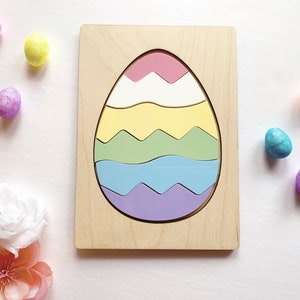 May include: A wooden puzzle board featuring a large egg-shaped cutout filled with five interlocking wavy pieces in pastel pink, white, yellow, light green, light blue, and lavender. Small decorative foam eggs and artificial flowers surround it.