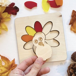 Pu&ograve; includere: Un puzzle di tacchino in legno con pezzi traslucidi colorati. Il tacchino ha un corpo rosso, arancione e giallo e una testa bianca con occhi neri. I pezzi del puzzle sono separati dal tabellone.
