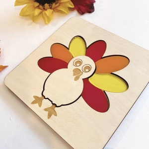 Pu&ograve; includere: Puzzle di legno a forma di tacchino con piume colorate. Il tacchino ha un corpo bianco, occhi neri e un becco rosso. Le piume sono gialle, arancioni e rosse.