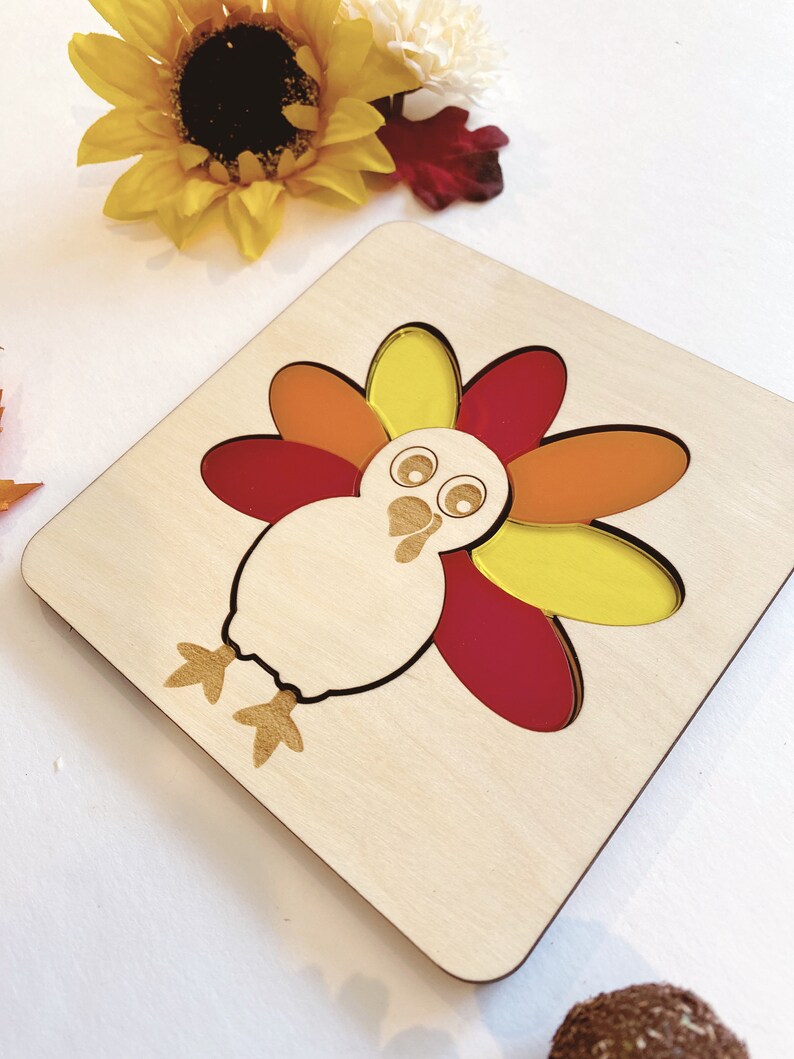Pu&ograve; includere: Puzzle di legno a forma di tacchino con piume colorate. Il tacchino ha un corpo bianco, occhi neri e un becco rosso. Le piume sono gialle, arancioni e rosse.