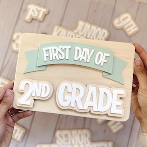 Peut inclure: Un panneau rectangulaire en bois avec une bannière vert clair portant le texte blanc FIRST DAY OF et le texte blanc 2ND GRADE en dessous. D'autres découpes en bois avec du texte comme 1ST, 9TH, JUNIOR et SENIOR KINDERGARTEN sont visibles.
