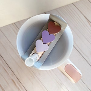 May include: A white mug with a pink handle and a straw. Three heart-shaped stickers in shades of purple and brown are attached to the mug.