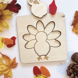 Pu&ograve; includere: Puzzle di legno a forma di tacchino con un ritaglio di tacchino e pezzi separati per le piume, la testa e la bargiglio. I pezzi sono rossi, arancioni e gialli.