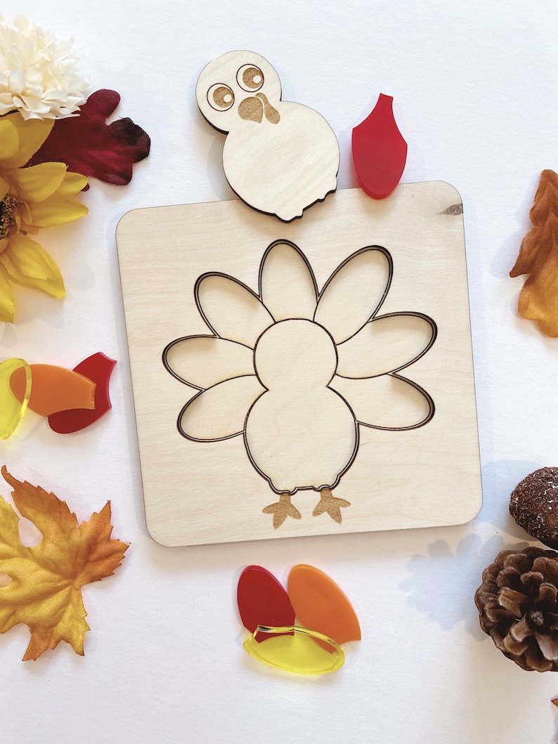 Pu&ograve; includere: Puzzle di legno a forma di tacchino con un ritaglio di tacchino e pezzi separati per le piume, la testa e la bargiglio. I pezzi sono rossi, arancioni e gialli.