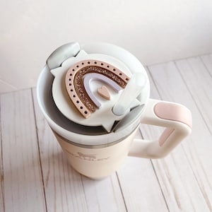 May include: A white travel mug with a pink handle and a lid featuring a rainbow and heart design. The mug has the word "Stanley" printed on the side.