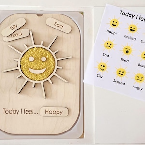 May include: A wooden board featuring a central yellow sun with a smiling face, surrounded by rays. It includes small wooden labels with emotions like Silly, Tired, Sad, and Happy. A matching paper chart displays sun icons with various facial expressions.