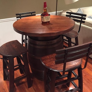 May include: A wooden table with a barrel base and a bottle of Maker's Mark bourbon whiskey on top. The table is surrounded by four black metal chairs with wooden seats.